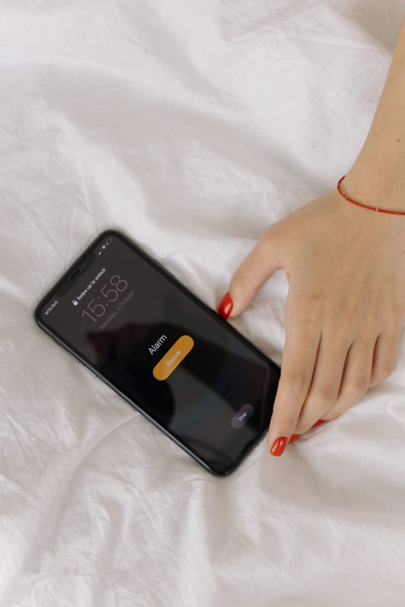 A close-up of a hand reaching to turn off an alarm on a smartphone resting on a white bed.